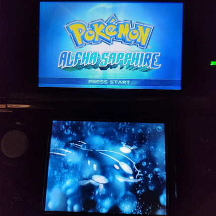A Nintendo DS handheld game console showing the Pokémon Alpha Sapphire game screen, with the blue game logo on the top display and an underwater scene on the bottom screen.