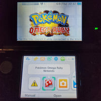 A Nintendo 3DS handheld console showing the Pokémon Omega Ruby game screen on the upper display and the app menu on the lower screen, photographed in a dark setting.