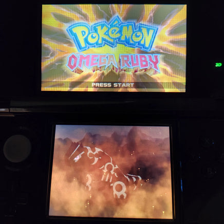 Nintendo 3DS handheld gaming console with the Pokémon Omega Ruby game cartridge inserted, showing the game's title screen on the upper screen and an in-game scene on the lower screen.