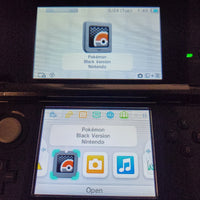 A close-up of a Nintendo 3DS handheld console showing the screen with a Pokémon app interface for the Black Version of the game. The top screen displays the Pokémon logo and text reading 'Pokémon Black Version Nintendo,' while the lower screen shows app icons including camera, music, and a highlighted Pokémon icon.