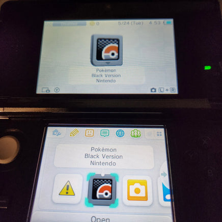 A close-up of a Nintendo DS handheld console showing the Pokémon Black Version game on the screen, with the top half displaying a Pokémon Black Version app icon and the bottom half showing app icons including a warning icon and camera icon.