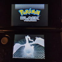 A black Nintendo DS handheld game console showing the Pokémon Black Version logo on the top screen and a white dragon character on the bottom screen, photographed in a dark setting.