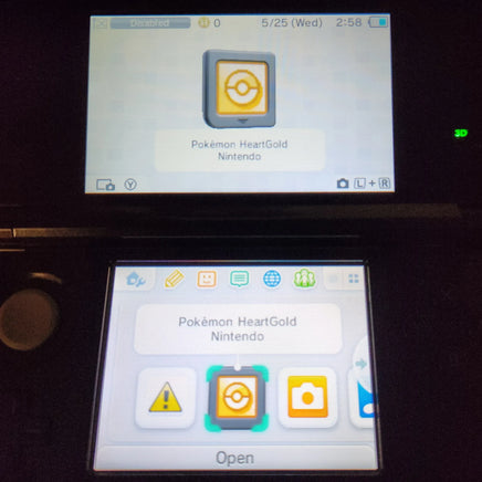 A Nintendo 3DS handheld console with two screens on, showing the Pokémon HeartGold app interface. The top screen displays the Pokémon HeartGold icon, and the bottom screen shows app icons including a warning icon, the HeartGold icon, a camera icon, and the word 'Open'.