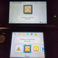 A close-up of a Nintendo 3DS handheld console showing the game menu for Pokémon HeartGold on both the upper screen and the lower screen.