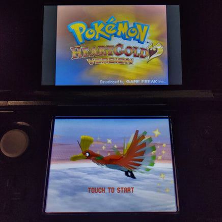 A Nintendo 3DS handheld game console displaying Pokémon HeartGold Version, with the main screen showing the game title and the lower screen showing a colorful bird character and the text 'TOUCH TO START'.