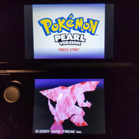 A Nintendo DS handheld game console showing the Pokémon Pearl Version title screen on the top display and a pink creature graphic on the bottom screen, photographed in a dark setting.