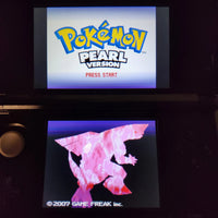 A Nintendo DS handheld gaming console showing the Pokémon Pearl Version title screen on the upper screen and a pink character on the lower screen, photographed in a dark setting.