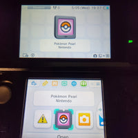A Nintendo 3DS handheld console showing the Pokémon Pearl Nintendo game icon on its screens, photographed in a dark setting.
