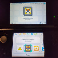 A Nintendo 3DS handheld console showing the Pokémon Platinum Version Nintendo app icon on its screens in a dark setting.
