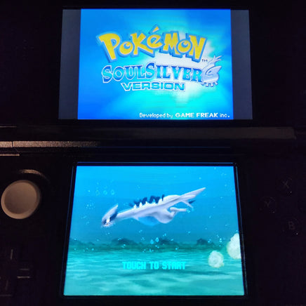 A Nintendo 3DS console showing the Pokémon SoulSilver Version game menu, with the top screen displaying the game title and the bottom screen showing a blue underwater scene with a white Pokémon and the text 'TOUCH TO START'.
