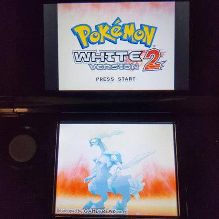 A Nintendo DS handheld game console with the Pokémon White 2 Version 2 game displayed on the screen, showing the game title on the top screen and a blue dragon character on the bottom screen.